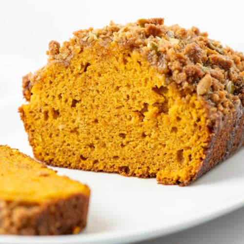 Side photo of a cut loaf of pumpkin bread with streusel topping so that you can see the inside with a slice of pumpkin bread on the side